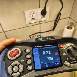 LOGOFORM Dariusz Dubrowski - Miernik instalacji elektrycznej CEM DT-6650 w dłoni, wyświetlający wyniki pomiarów gniazdka elektrycznego na tle płytek. Widoczny kabel zasilający.