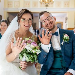 Dawid Czub - Uśmiechnięta para młoda w kościele prezentuje obrączki, ona z bukietem, on w eleganckim garniturze w kratę, w tle goście weselni.