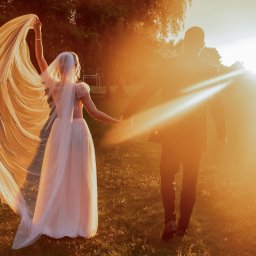 Dawid Czub - Para młoda spaceruje trzymając się za ręce w promieniach zachodzącego słońca. Panna młoda trzyma welon. Ujęcie zza pleców, ciepłe światło.