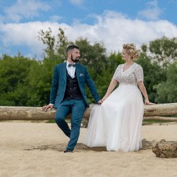 Dawid Czub - Para młoda w plenerze: on w turkusowym garniturze, ona w koronkowej sukni, siedzą na pniu drzewa na piaszczystej plaży, zieleń w tle, błękitne niebo z chmurami.