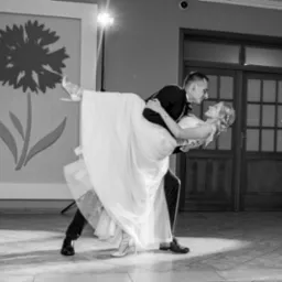 Czarno-białe zdjęcie pary młodej w tańcu, panna młoda w sukni uniesiona przez pana młodego. W tle dekoracja z motywem kwiatowym i światło reflektora.