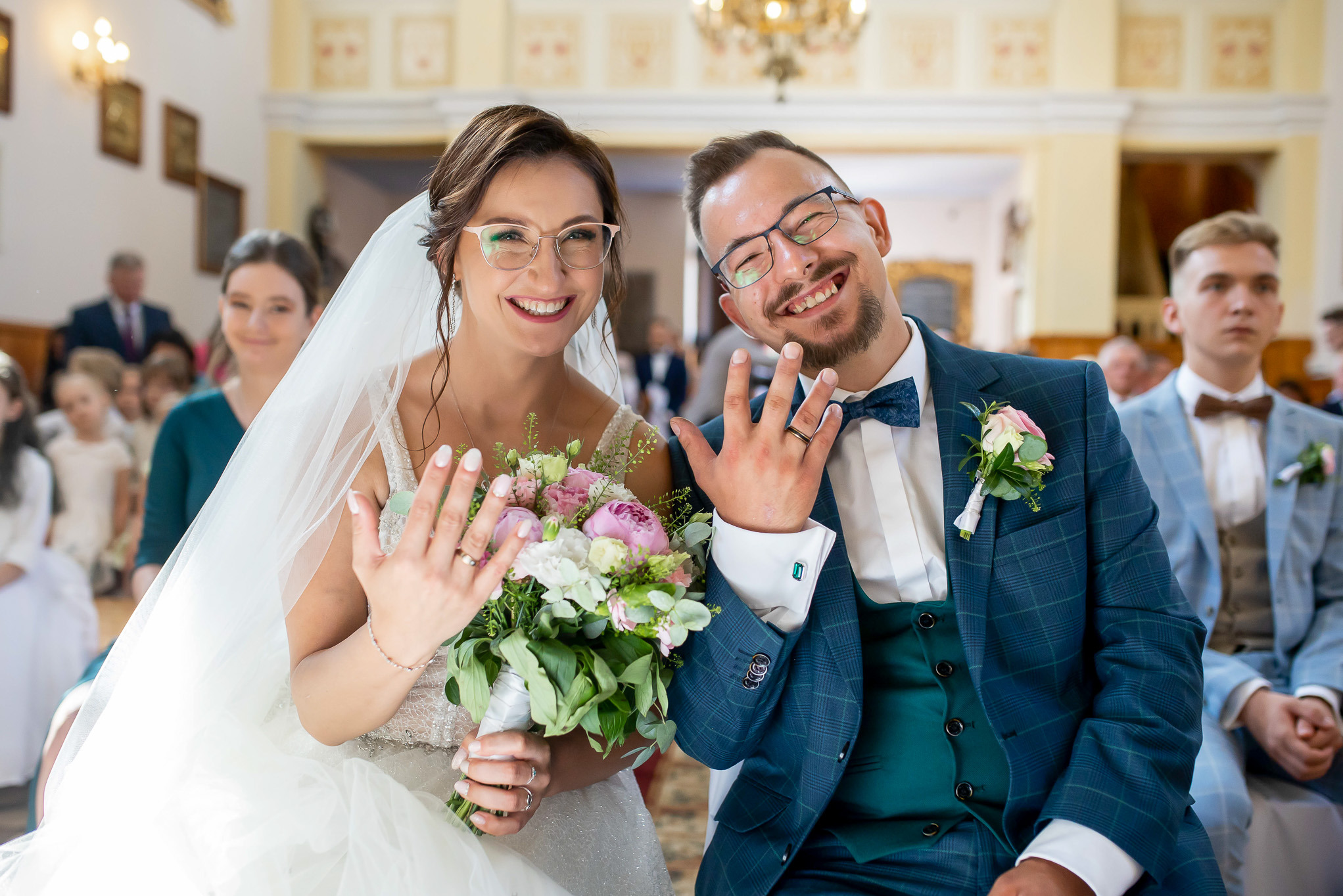 Uśmiechnięta para młoda w kościele prezentuje obrączki, ona z bukietem, on w eleganckim garniturze w kratę, w tle goście weselni.