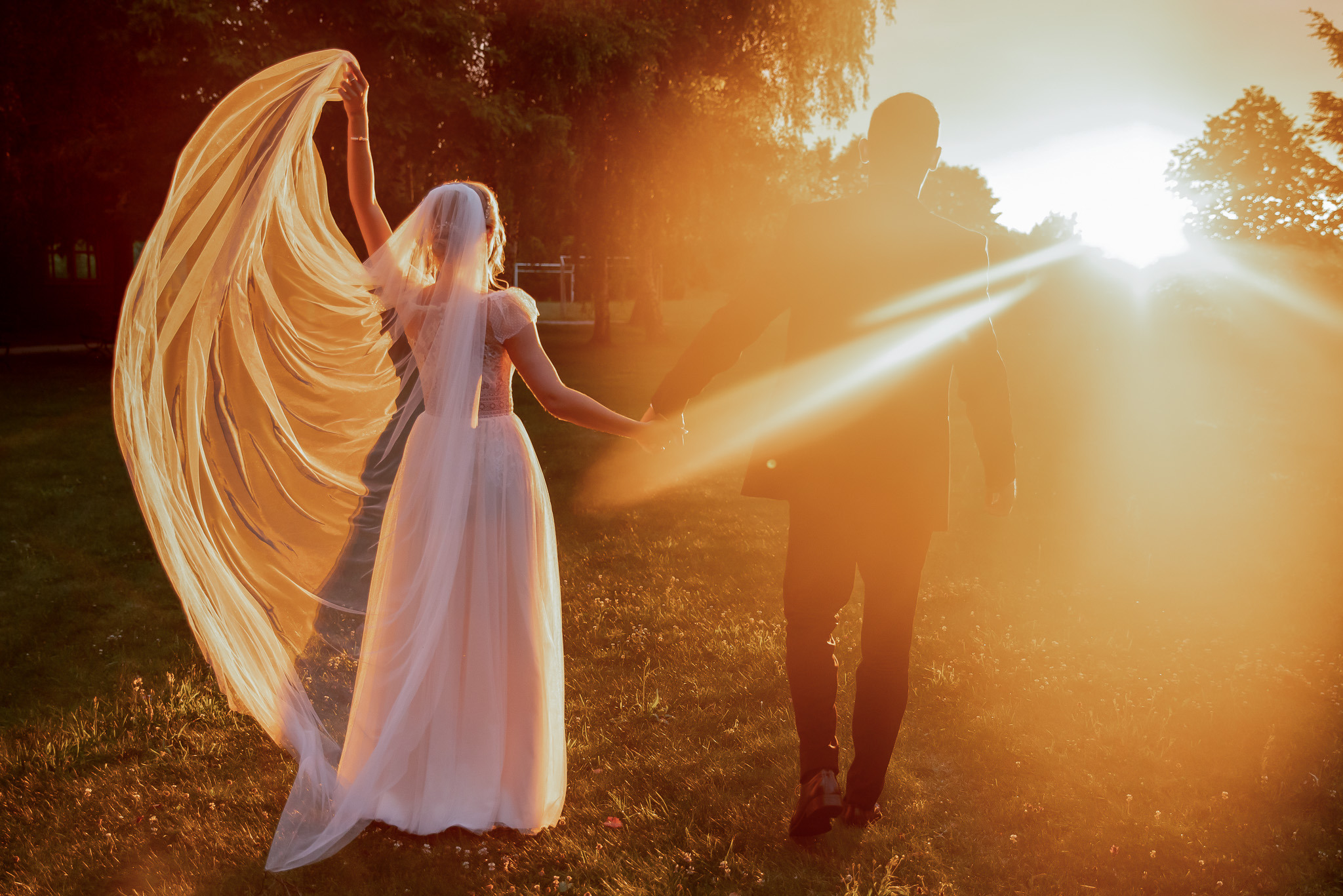 Para młoda spaceruje trzymając się za ręce w promieniach zachodzącego słońca. Panna młoda trzyma welon. Ujęcie zza pleców, ciepłe światło.