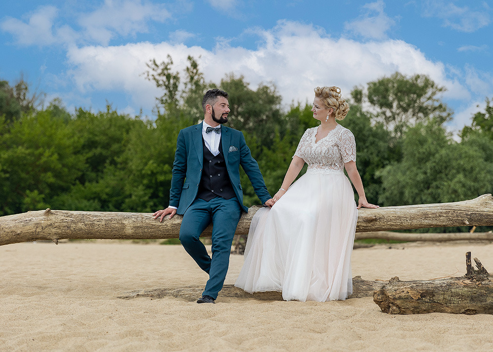 Para młoda w plenerze: on w turkusowym garniturze, ona w koronkowej sukni, siedzą na pniu drzewa na piaszczystej plaży, zieleń w tle, błękitne niebo z chmurami.