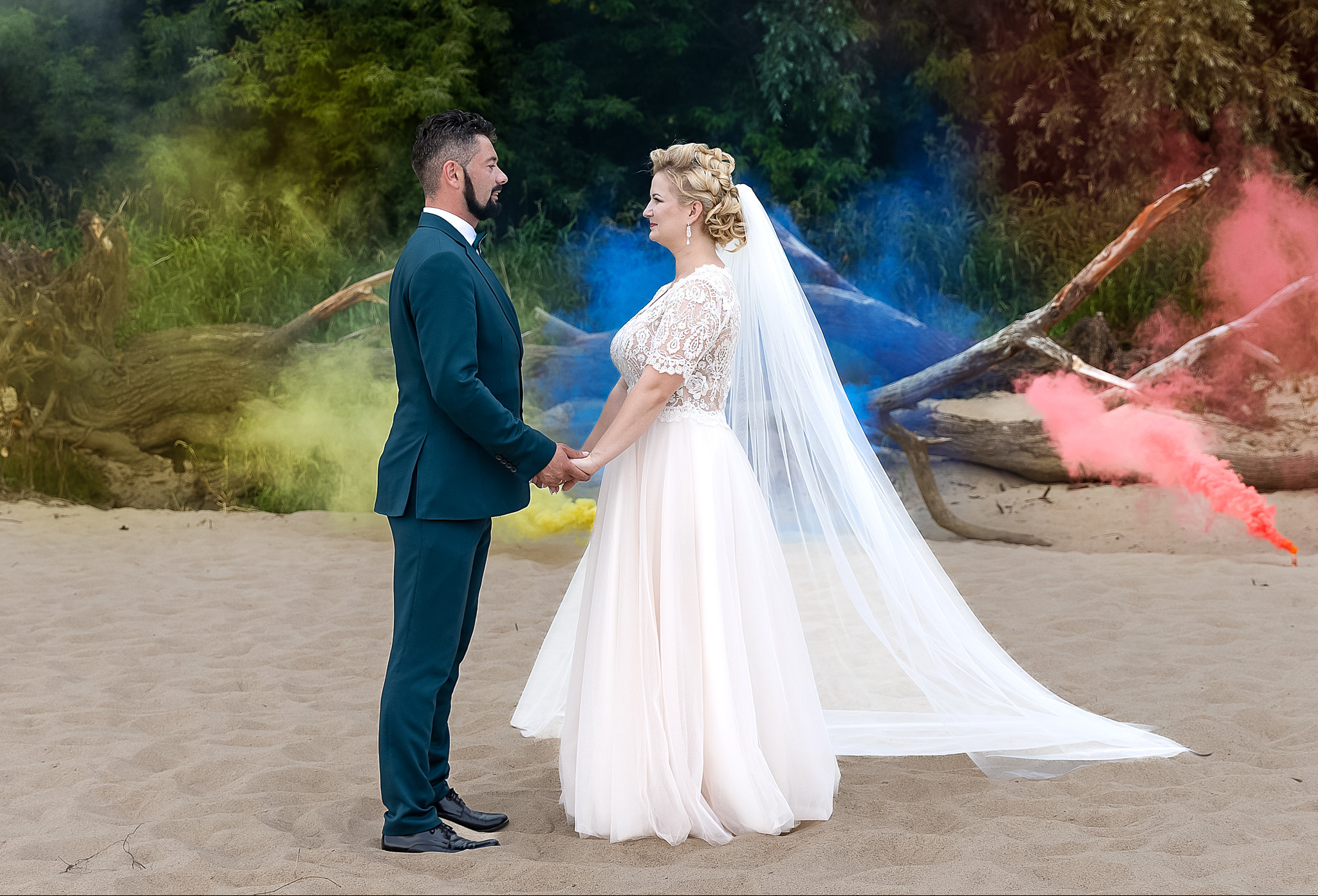 Para młoda na plaży trzyma się za ręce, otoczona kolorowymi dymami i naturalnym tłem z zieleni i drewna. Pan młody w ciemnozielonym garniturze, panna młoda w koronkowej sukni.