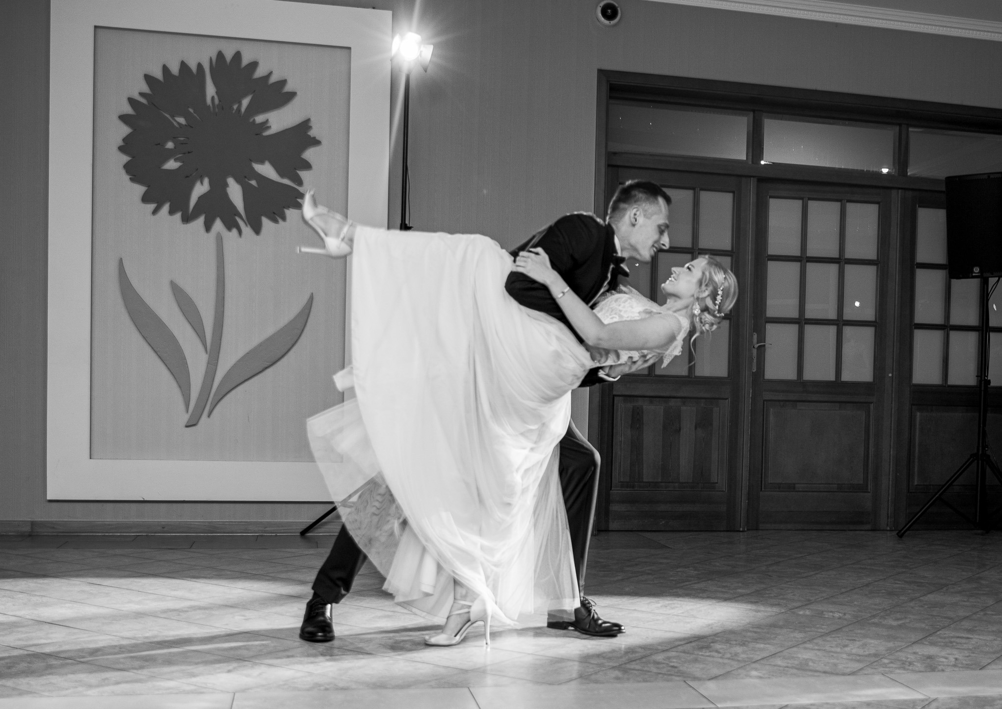 Czarno-białe zdjęcie pary młodej w tańcu, panna młoda w sukni uniesiona przez pana młodego. W tle dekoracja z motywem kwiatowym i światło reflektora.