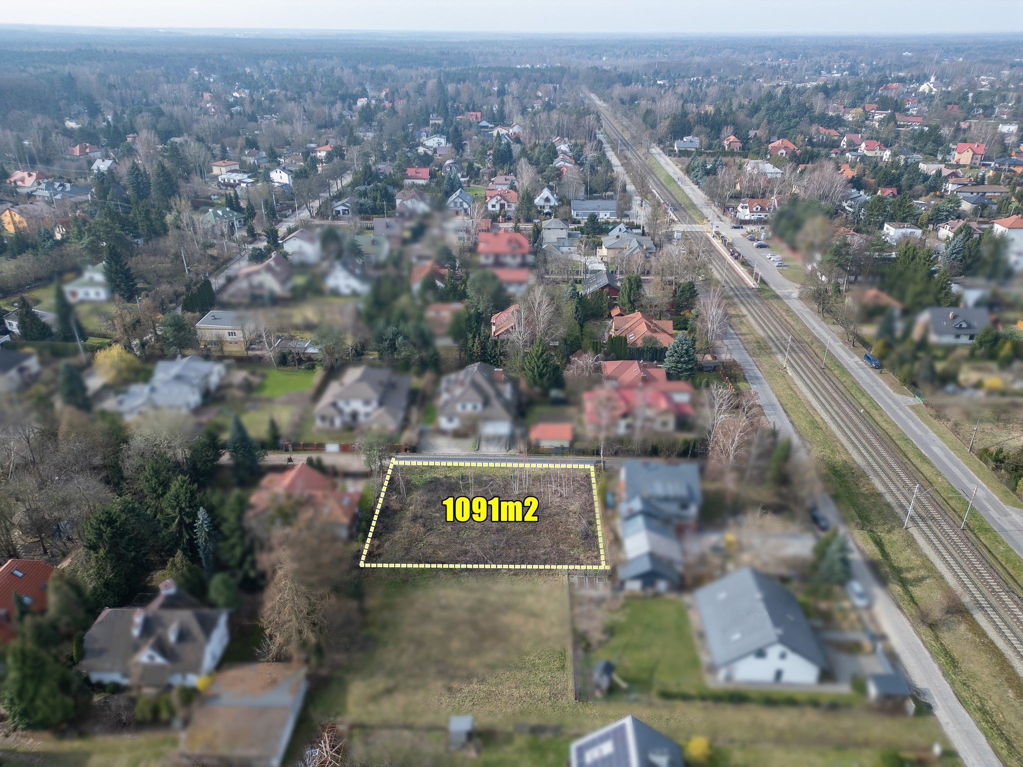 Zdjęcie z drona przedstawia działkę budowlaną o powierzchni 1091m2, otoczoną zabudową jednorodzinną i linią kolejową w tle. Działka oznaczona żółtą linią.