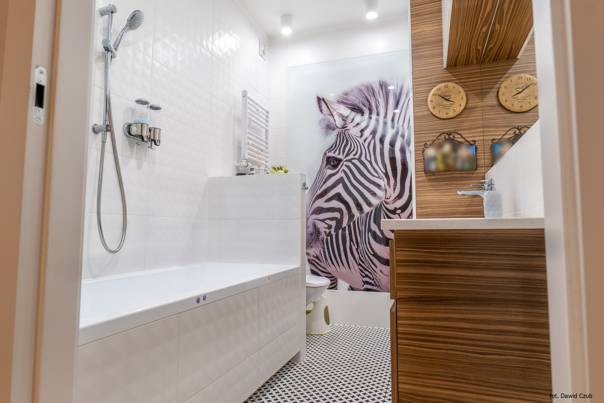 Nowoczesna łazienka z białą wanną, mozaikową podłogą i akcentem w postaci grafiki zebry na ścianie. Drewniana szafka pod umywalką z oryginalnymi zegarami.
