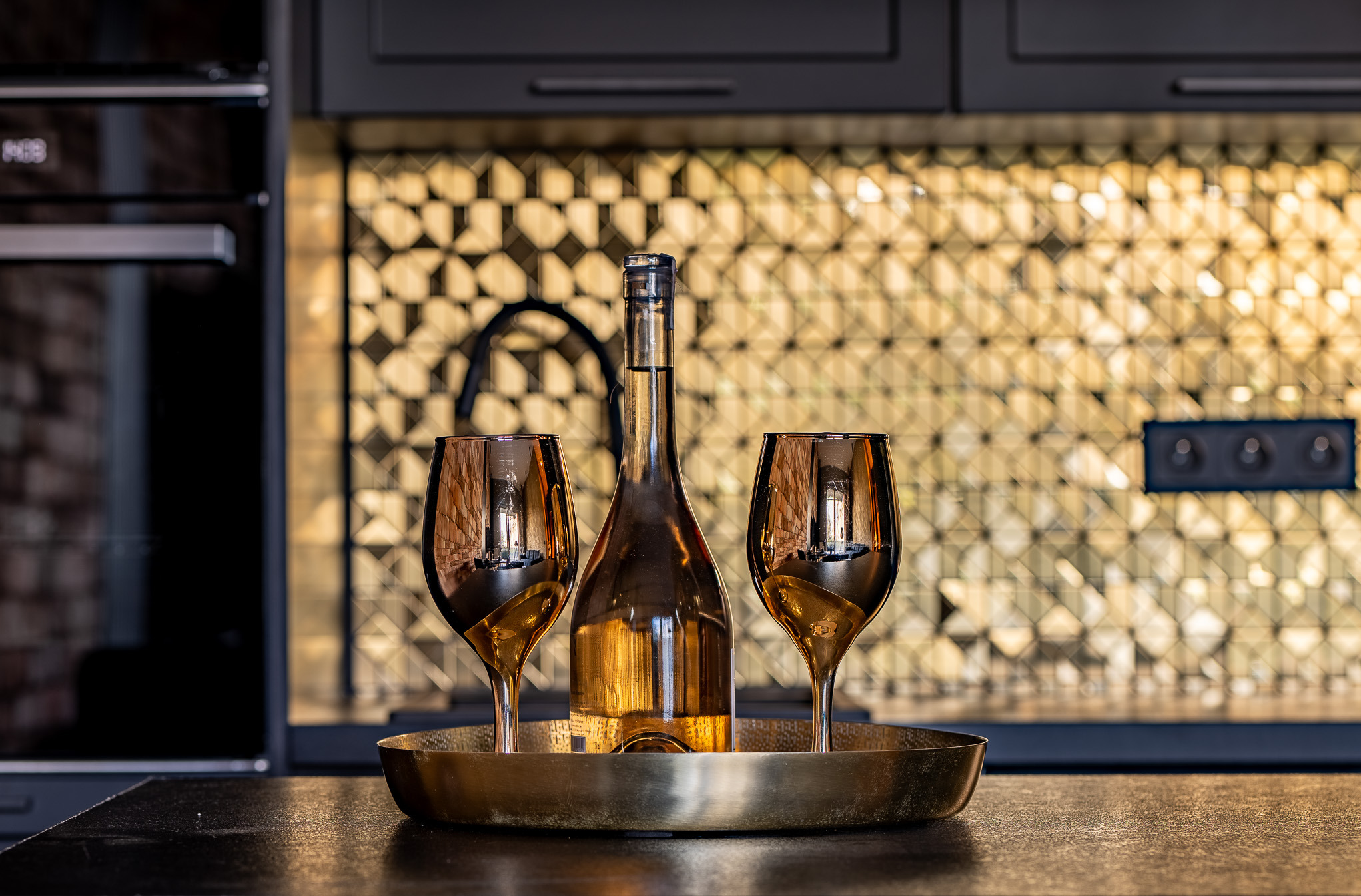 Butelka z bursztynowym trunkiem i dwa kieliszki na złotej tacy. W tle mozaika w geometryczne wzory i czarne szafki kuchenne.