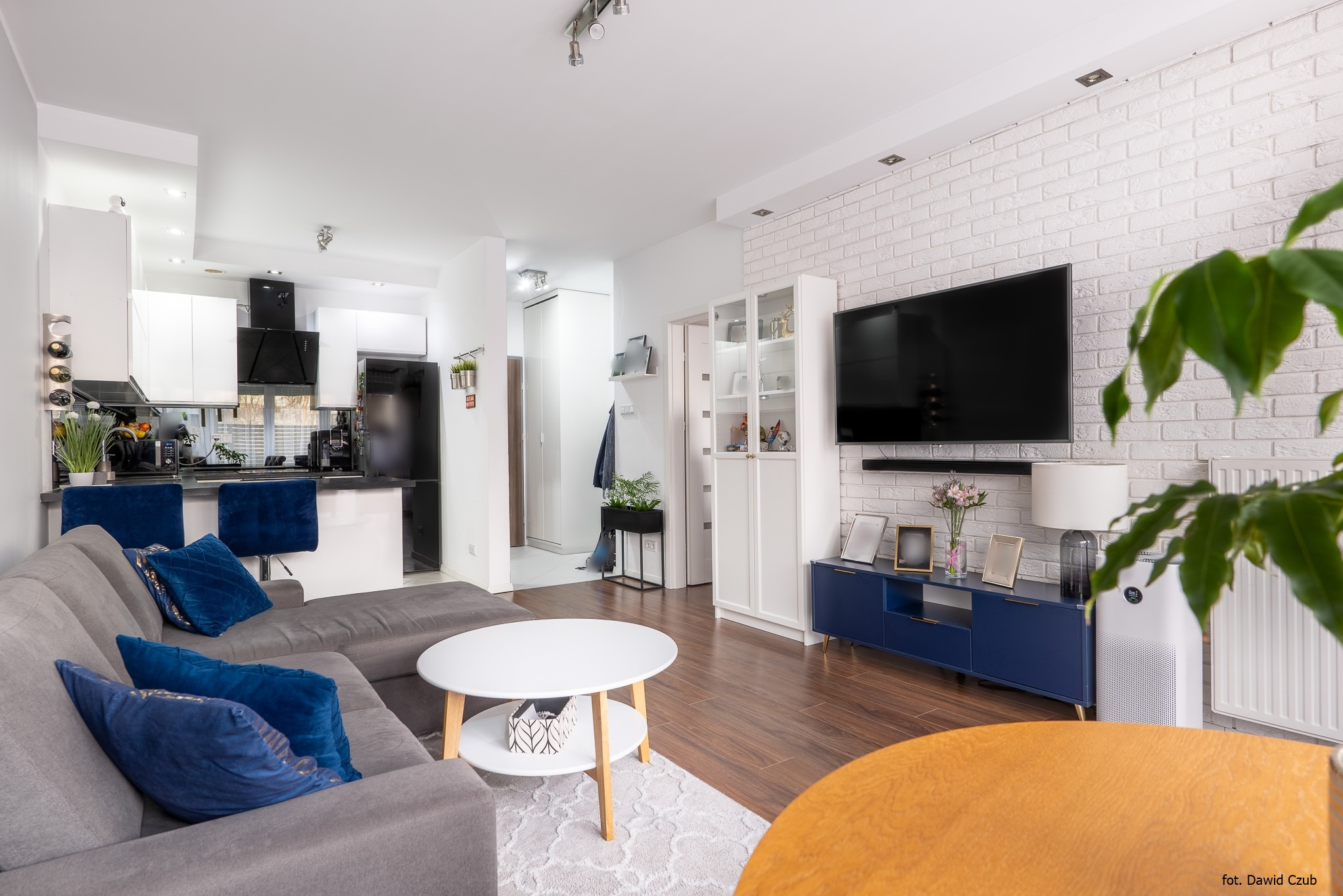 Jasne wnętrze: salon z aneksem kuchennym, szara sofa z niebieskimi poduszkami, biały stolik kawowy i ceglana ściana z telewizorem. Nowoczesny design.