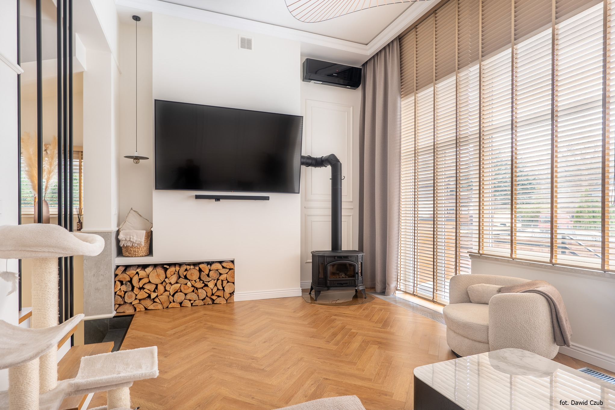 Jasne wnętrze z parkietem w jodełkę, kominkiem, telewizorem i drewnianymi żaluzjami. W rogu stoi beżowy fotel i stolik kawowy.