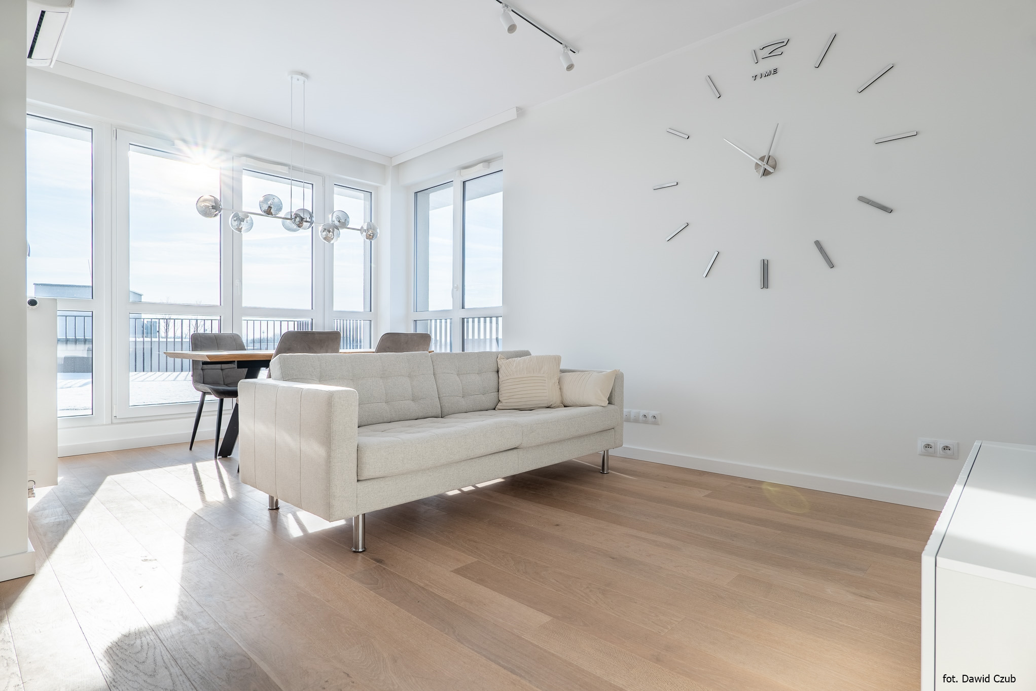 Jasny salon z parkietem, beżową sofą i dużym zegarem na ścianie. Okna wpuszczają dużo światła, oświetlając stół z krzesłami.