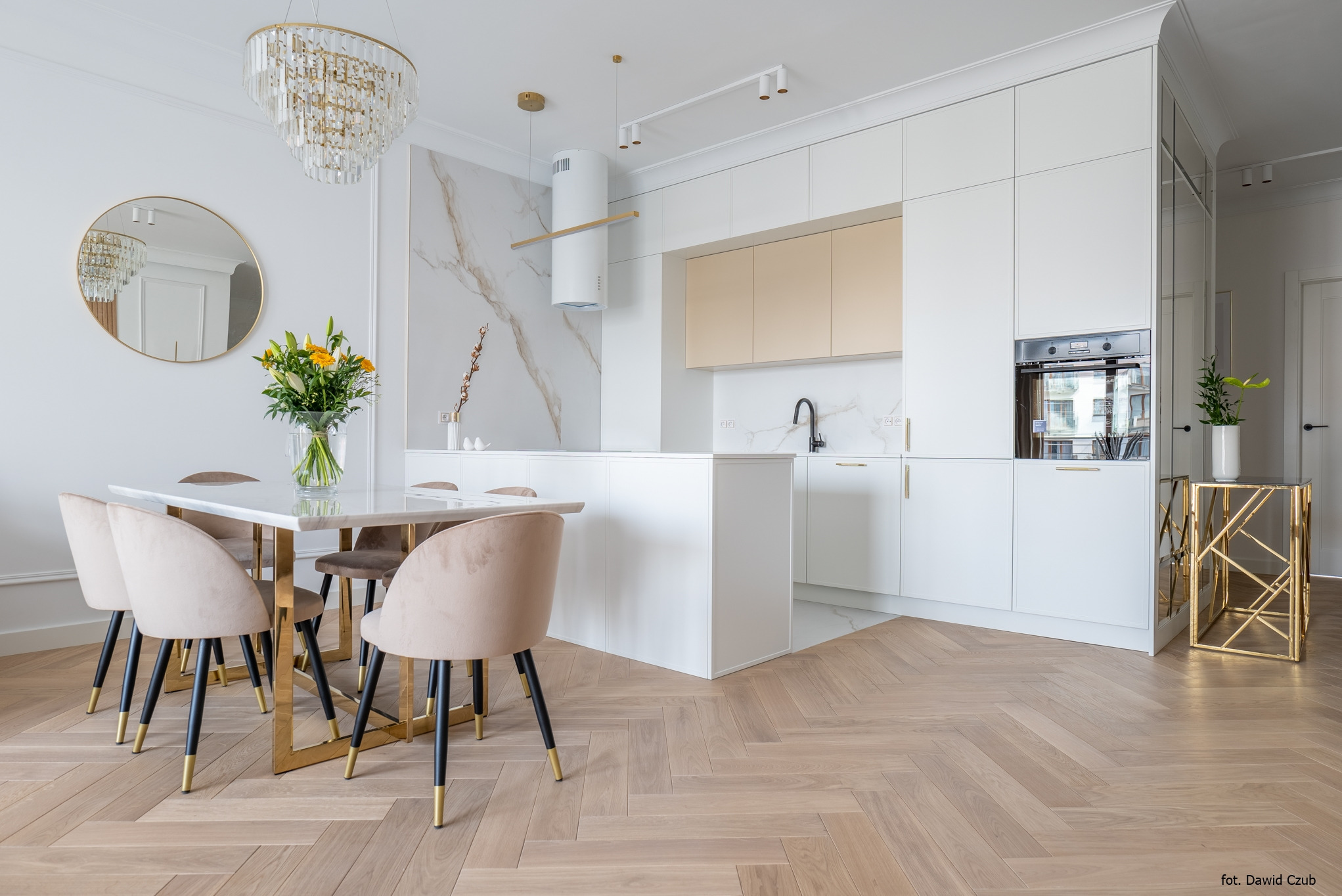 Jasna kuchnia z wyspą i marmurowym blatem, stół z kwiatami, parkiet w jodełkę. Nowoczesne oświetlenie, złote akcenty. Fotografia wnętrz.