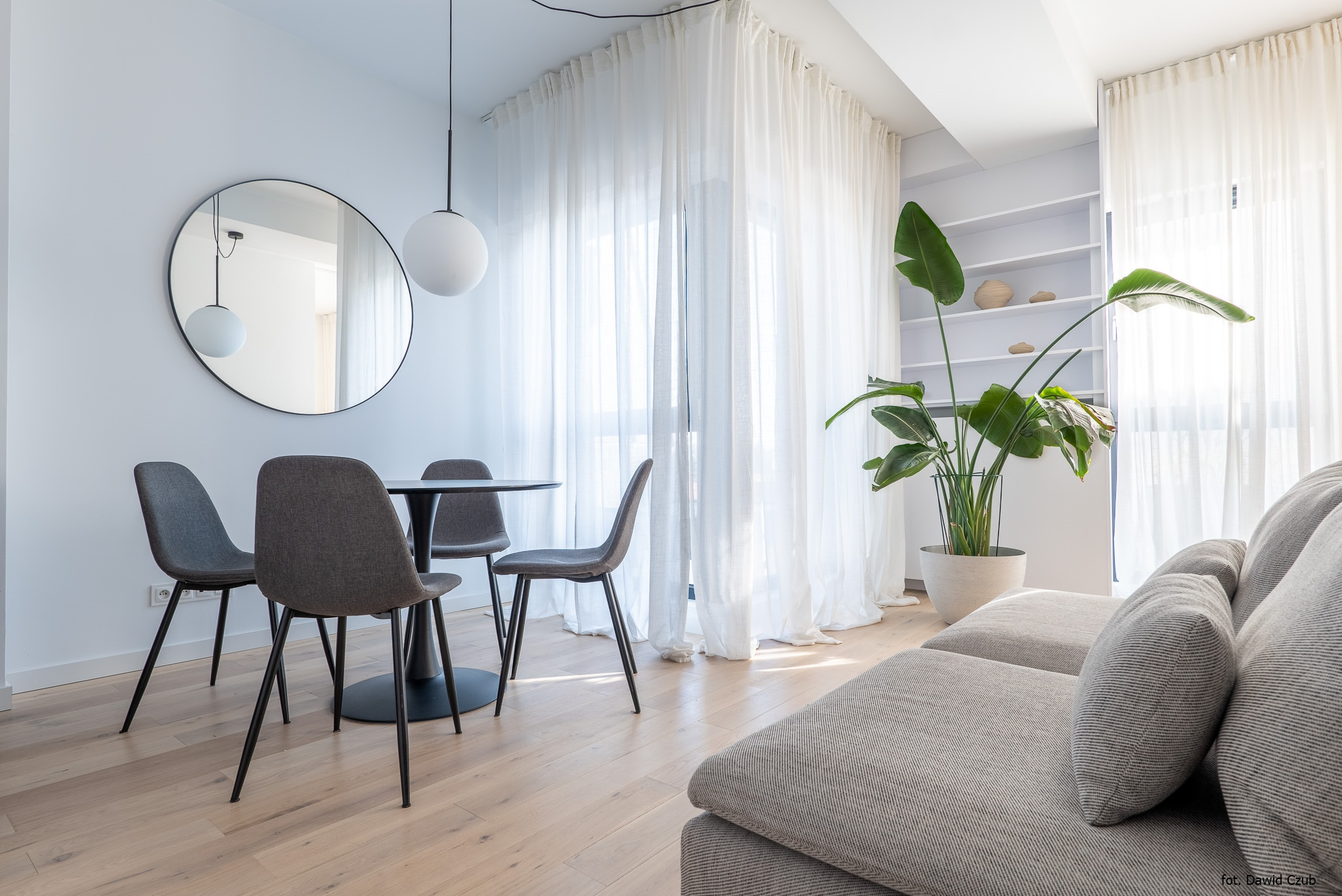 Jasne wnętrze: okrągły stół z krzesłami, okrągłe lustro, lampa kula, sofa i roślina w doniczce. Minimalistyczny design, naturalne światło.