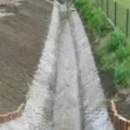 Wykończony drenaż liniowy z ażurowych płyt betonowych, obramowany niskim płotkiem z drewna, widok z góry na tle trawnika i ogrodzenia.