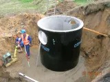 Studnia kanalizacyjna z betonu w wykopie, widoczne otwory boczne, piasek i pracownicy w tle. Ujęcie z góry, z bliska, w dziennym oświetleniu.