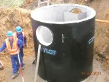 Czarna studzienka kanalizacyjna z otworem w ziemi, widoczni robotnicy w kaskach i kamizelkach odblaskowych. Etap prac instalacyjnych.