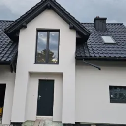 Elewacja domu jednorodzinnego z czarną stolarką okienną i grafitową dachówką, widok na front budynku z garażem w trakcie prac wykończeniowych.