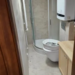 Mała łazienka po remoncie: kabina prysznicowa, bojler, wisząca toaleta z umywalką i szafka pod umywalkę w jasnych kolorach. Widok zza otwartych drzwi.