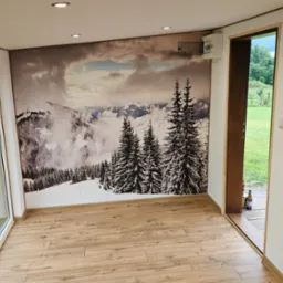 Wnętrze pokoju z fototapetą górskiego krajobrazu. Jasne ściany, drewniana podłoga, okno z widokiem na zieleń. Uwagę zwraca fototapeta z motywem gór.