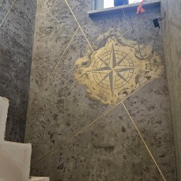 Bo Bau - Dekoracyjna ściana z efektem betonu i złotymi liniami, kompas na ścianie, schody w trakcie wykończenia, surowe wnętrze z oknem.