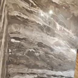 Ściana z dekoracyjną, szarą farbą imitującą marmur z delikatnymi, złotymi żyłkami. Widoczny narożnik i zabezpieczenie podłogi folią malarską.