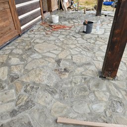 Bo Bau - Ukończona kamienna posadzka na zewnątrz budynku z drewnianymi elementami konstrukcyjnymi i widocznymi narzędziami pracy, z bliska, z detalami faktury kamienia.