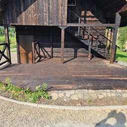 Bo Bau - Ciemna, drewniana altana z schodami i podestem, obrzeża z kamienia. Widoczne zarośla i cień osoby.