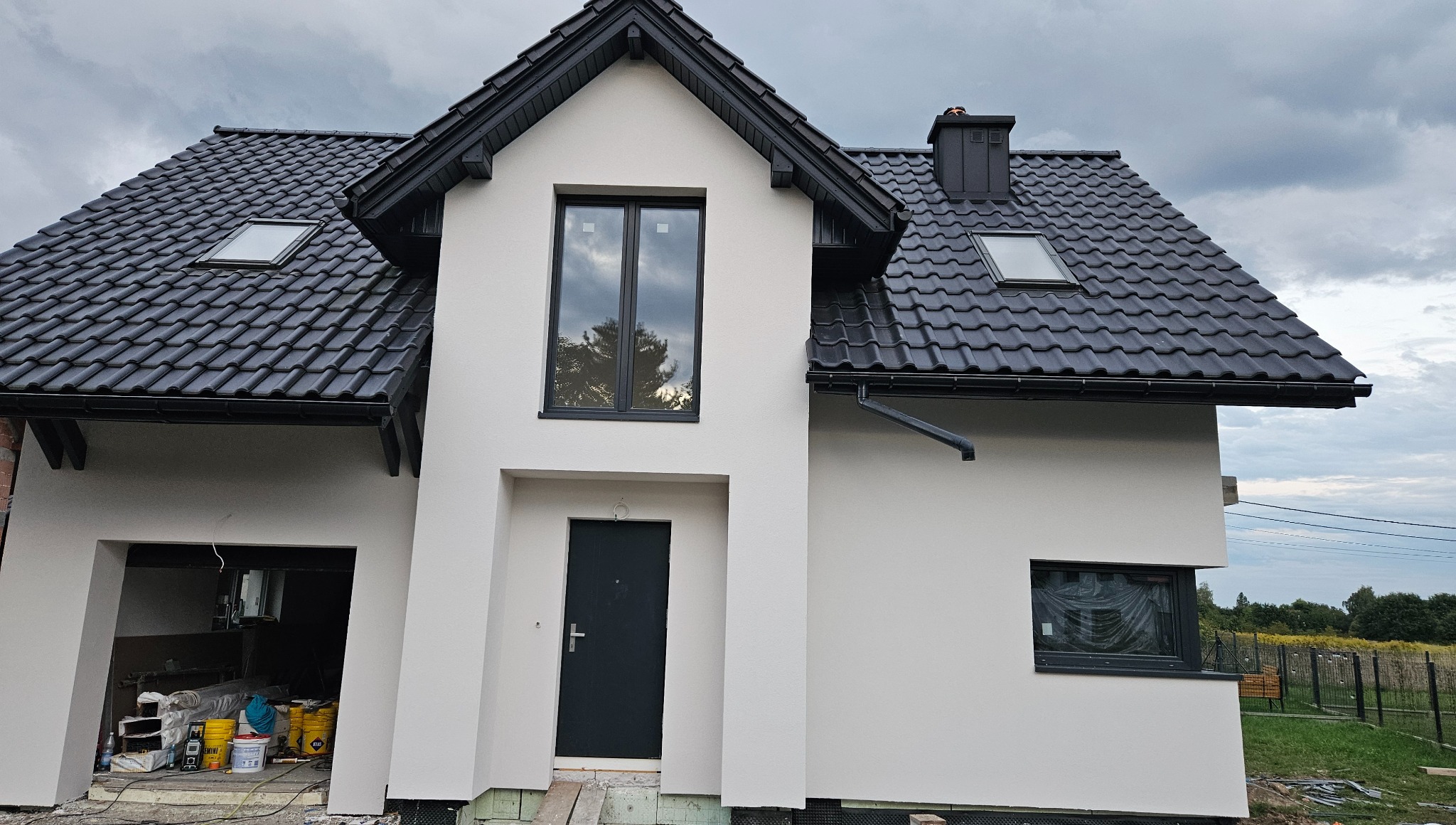 Elewacja domu jednorodzinnego z czarną stolarką okienną i grafitową dachówką, widok na front budynku z garażem w trakcie prac wykończeniowych.