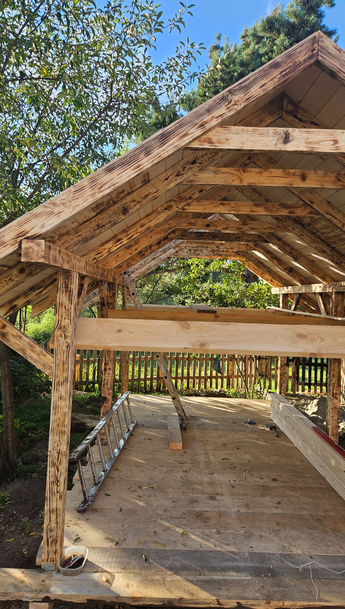 Konstrukcja altany w trakcie budowy, widoczne drewniane elementy konstrukcyjne, drabina i narzędzia. Otoczenie zielone, słoneczny dzień.