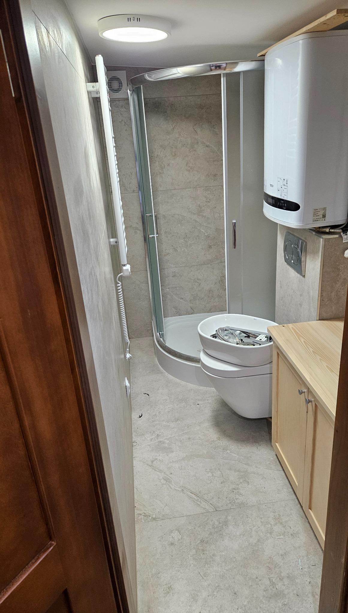 Mała łazienka po remoncie: kabina prysznicowa, bojler, wisząca toaleta z umywalką i szafka pod umywalkę w jasnych kolorach. Widok zza otwartych drzwi.