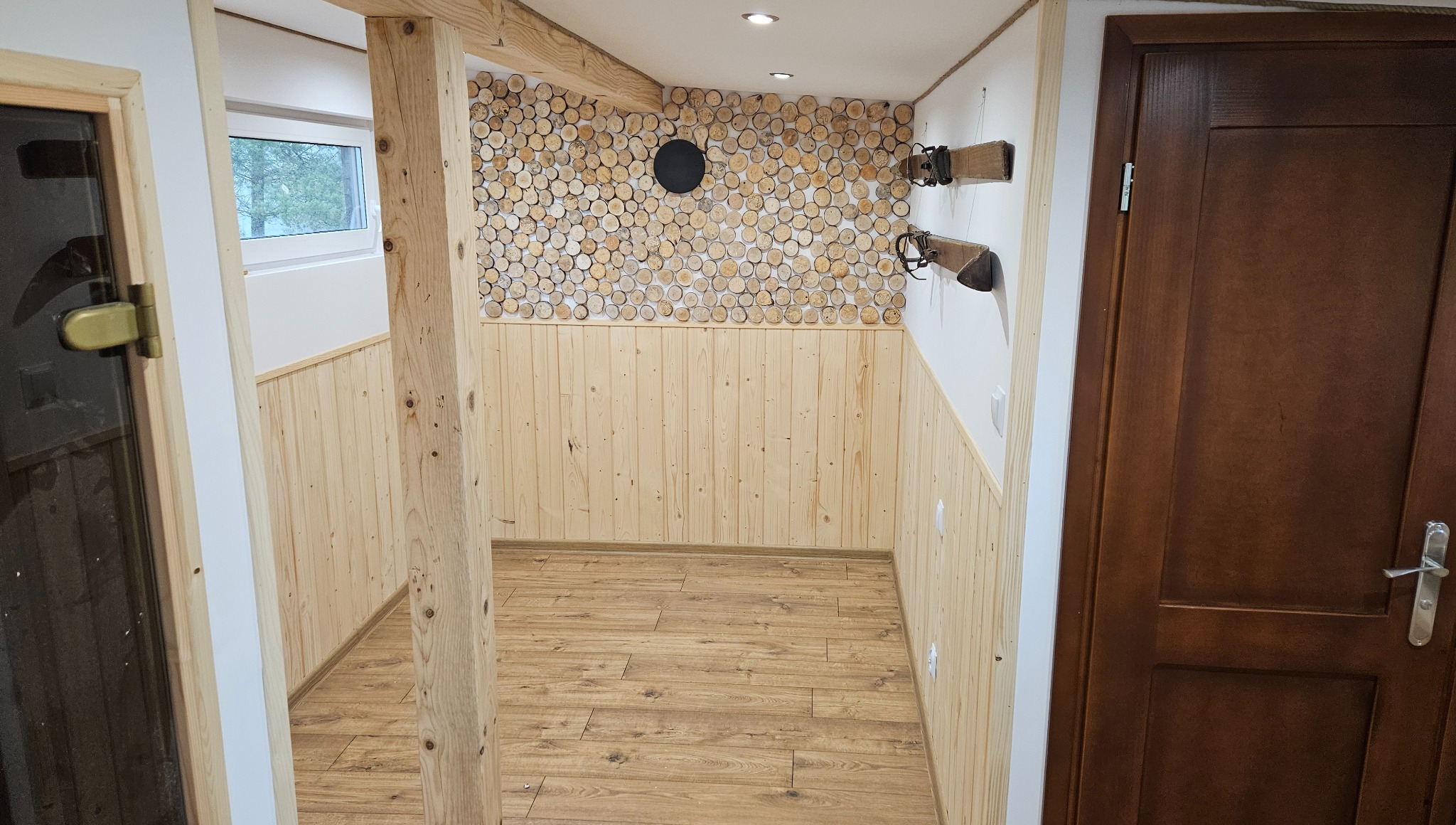 Drewniane wykończenie wnętrza: boazeria, podłoga z paneli, ściana z drewnianych krążków. Widoczne drzwi do sauny i ciemne drzwi po prawej.