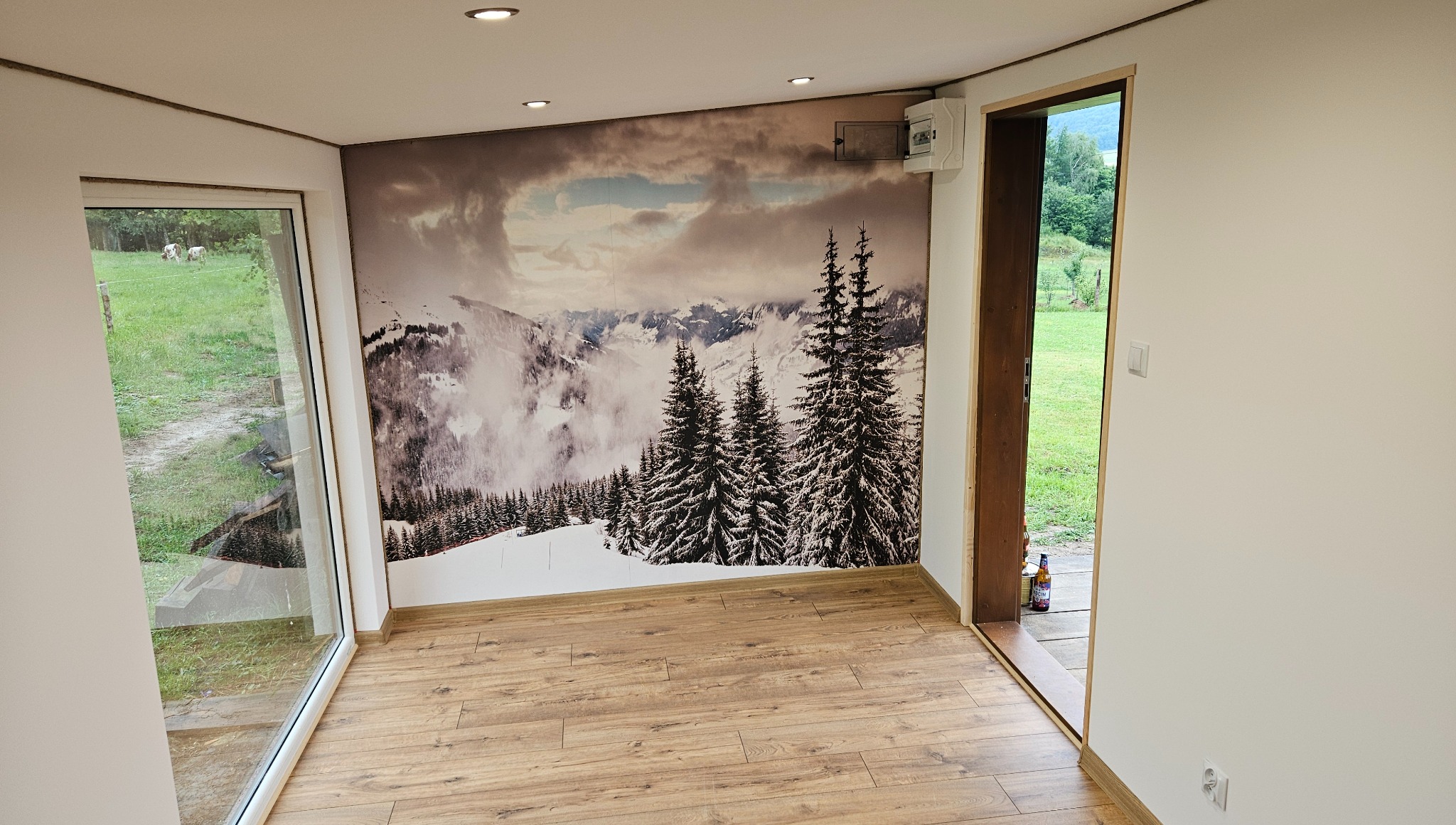 Wnętrze pokoju z fototapetą górskiego krajobrazu. Jasne ściany, drewniana podłoga, okno z widokiem na zieleń. Uwagę zwraca fototapeta z motywem gór.