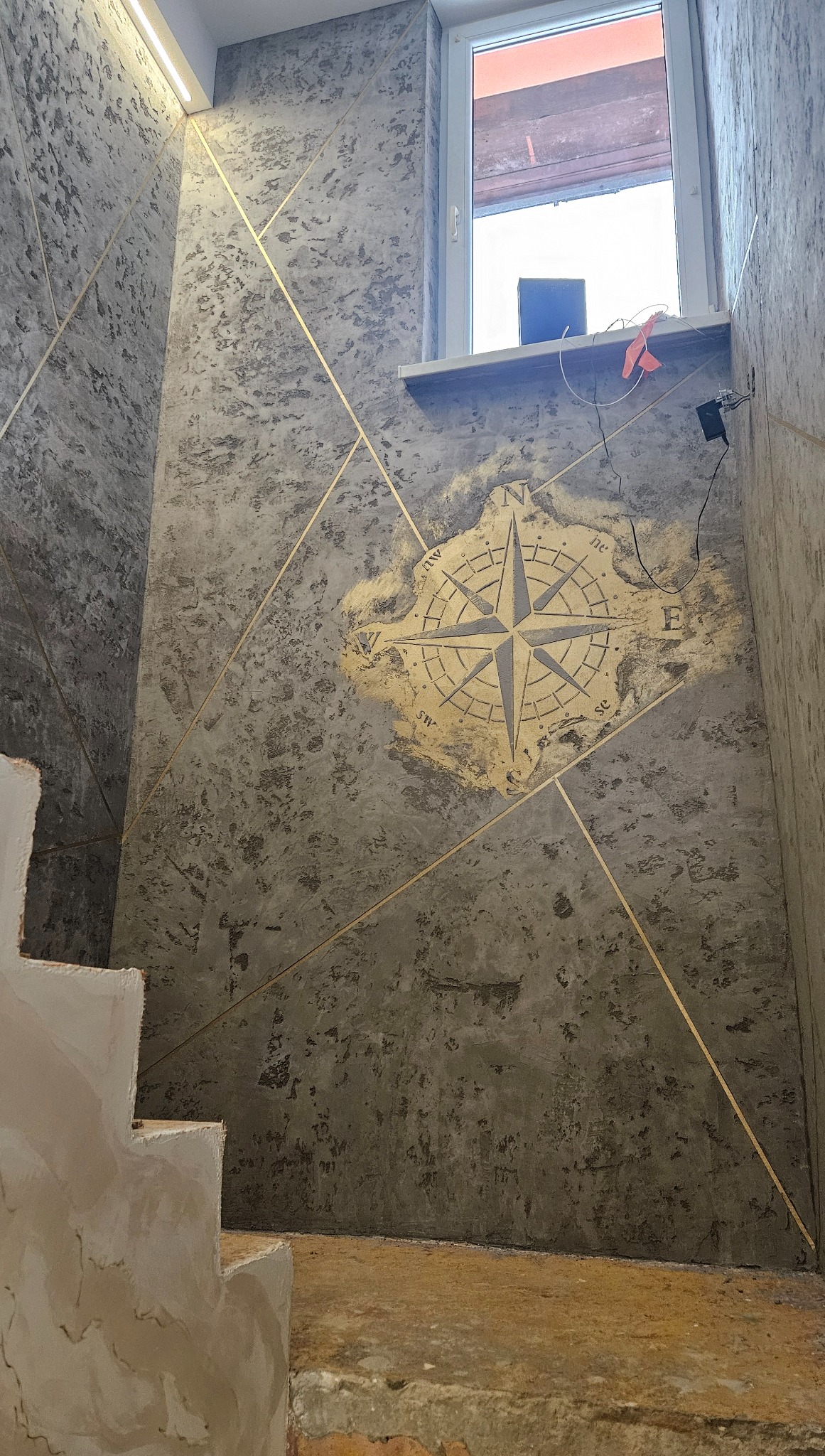 Dekoracyjna ściana z efektem betonu i złotymi liniami, kompas na ścianie, schody w trakcie wykończenia, surowe wnętrze z oknem.