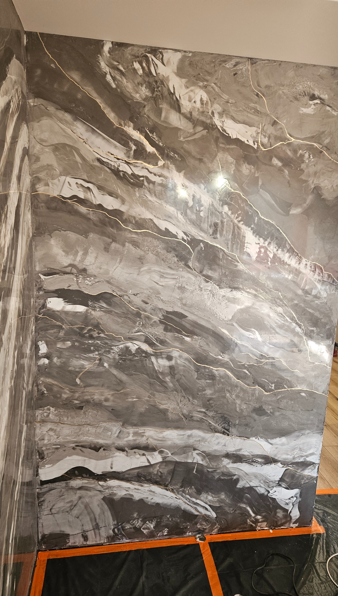 Ściana z dekoracyjną, szarą farbą imitującą marmur z delikatnymi, złotymi żyłkami. Widoczny narożnik i zabezpieczenie podłogi folią malarską.