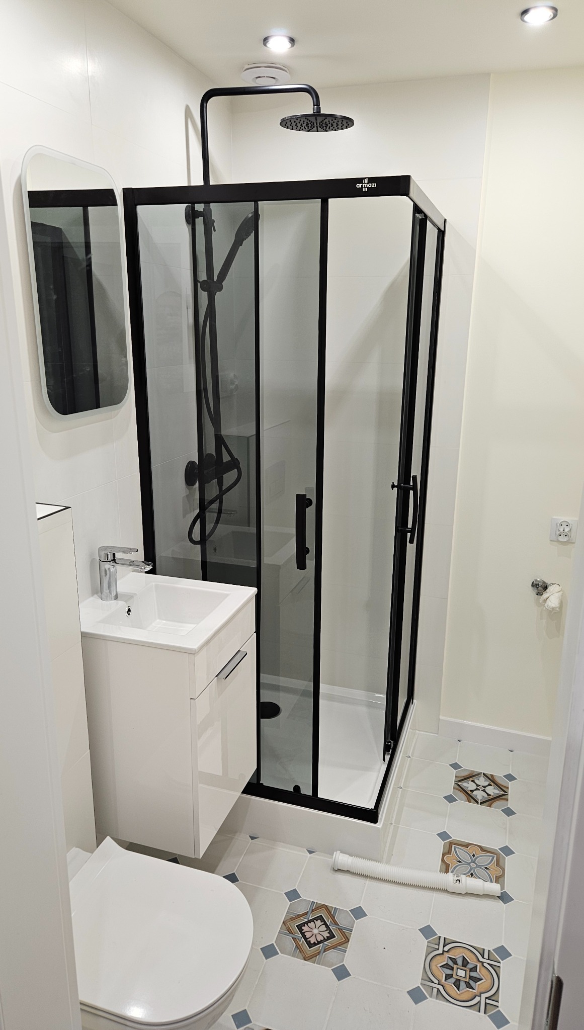 Nowoczesna łazienka po remoncie: kabina prysznicowa z czarnymi ramami, biała umywalka z szafką, toaleta i dekoracyjne płytki podłogowe z wzorem.