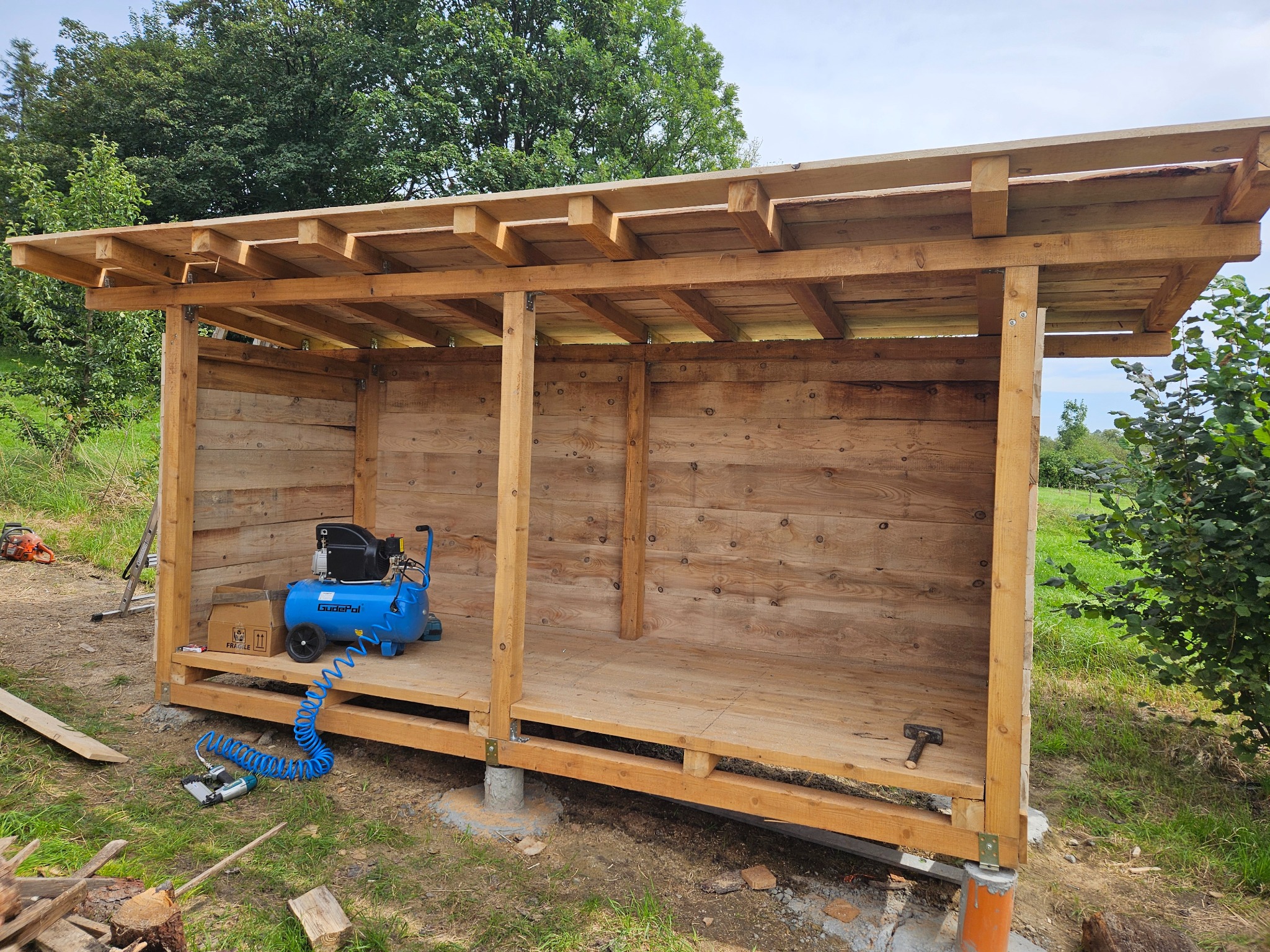 Drewniana konstrukcja altany w trakcie budowy, widoczny kompresor i narzędzia. Konstrukcja oparta na betonowych fundamentach, w tle zieleń.