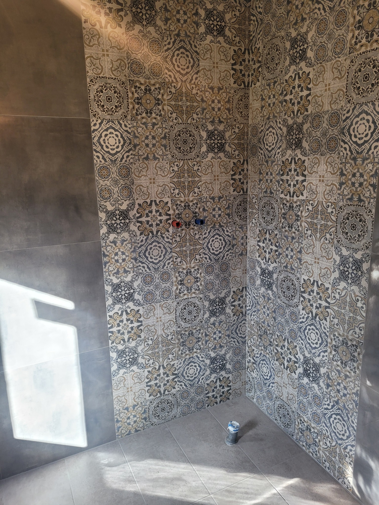 Wykończona wnęka prysznicowa z dekoracyjnymi płytkami patchwork w odcieniach beżu i szarości, widoczne przyłącza wodne i odpływ podłogowy.