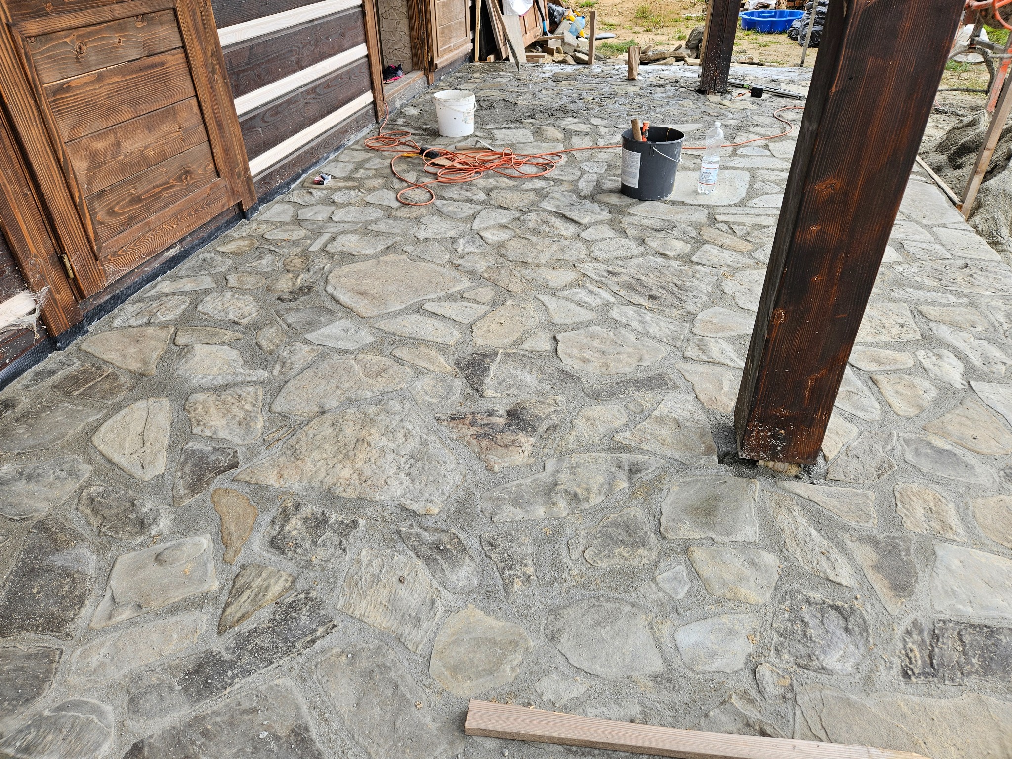 Ukończona kamienna posadzka na zewnątrz budynku z drewnianymi elementami konstrukcyjnymi i widocznymi narzędziami pracy, z bliska, z detalami faktury kamienia.