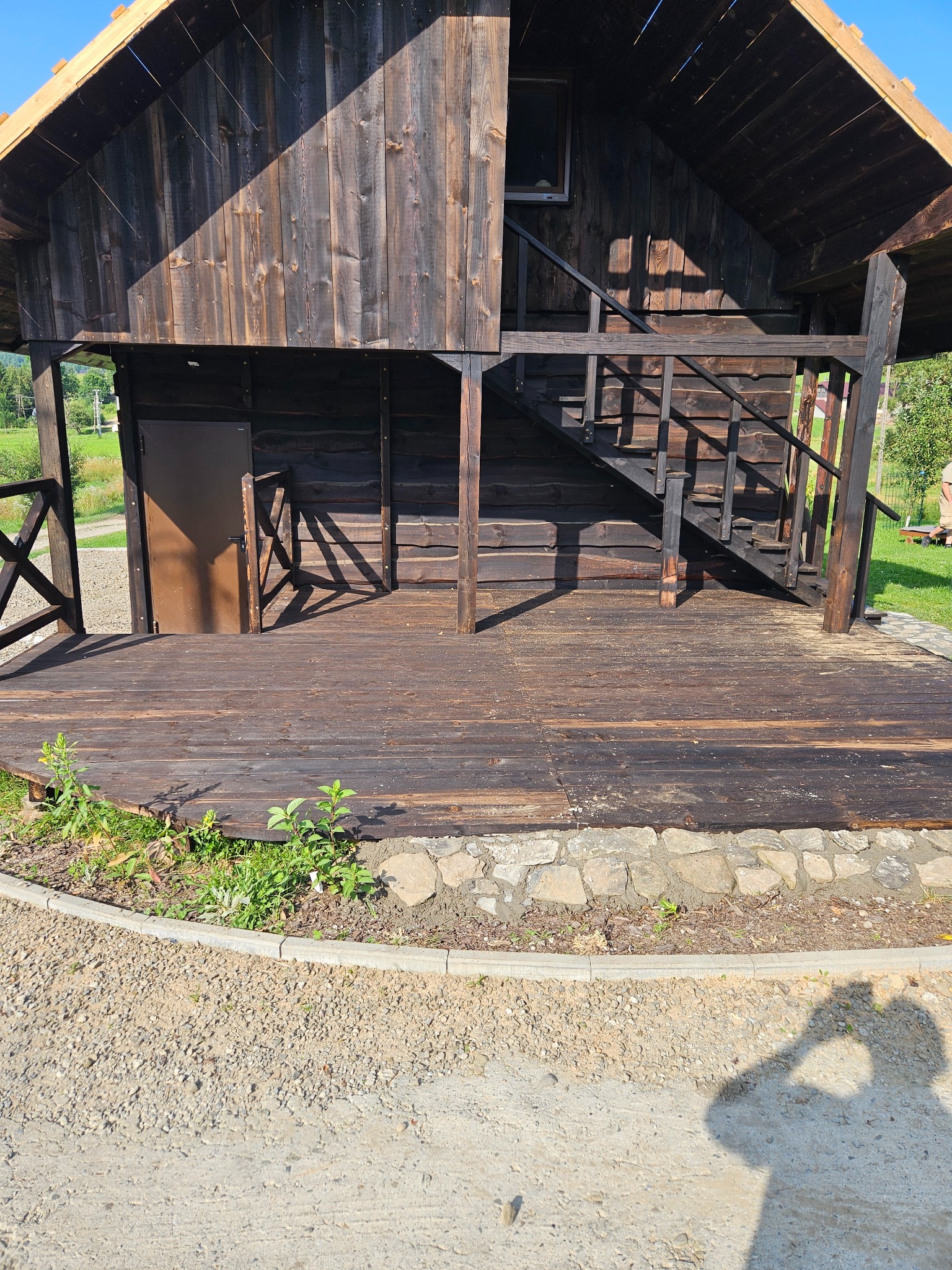 Ciemna, drewniana altana z schodami i podestem, obrzeża z kamienia. Widoczne zarośla i cień osoby.