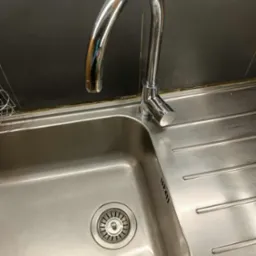 Błyszczący kran nad czystym, metalowym zlewem kuchennym z odpływem. Widoczne ślady po czyszczeniu, ciemna ściana w tle. Ujęcie z góry, ukośne.