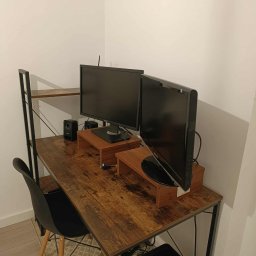 Czystonauta - Paweł Urbański - Biurko z dwoma monitorami na drewnianych podstawkach, czarne głośniki i krzesło. Widoczne kable pod biurkiem i kosz na śmieci. Minimalistyczny styl, jasne ściany.