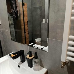 Czystonauta - Paweł Urbański - Szara łazienka po remoncie: umywalka z czarną armaturą, dozowniki mydła, lustro odbijające toaletę i grzejnik. Nowoczesne płytki, minimalistyczny design.