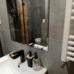 Szara łazienka po remoncie: umywalka z czarną armaturą, dozowniki mydła, lustro odbijające toaletę i grzejnik. Nowoczesne płytki, minimalistyczny design.