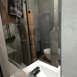 Czystonauta - Paweł Urbański - Nowoczesna łazienka z szarymi płytkami, kabiną prysznicową i białym umywalką, widziana w odbiciu lustra. Widoczne ręczniki i akcesoria łazienkowe.