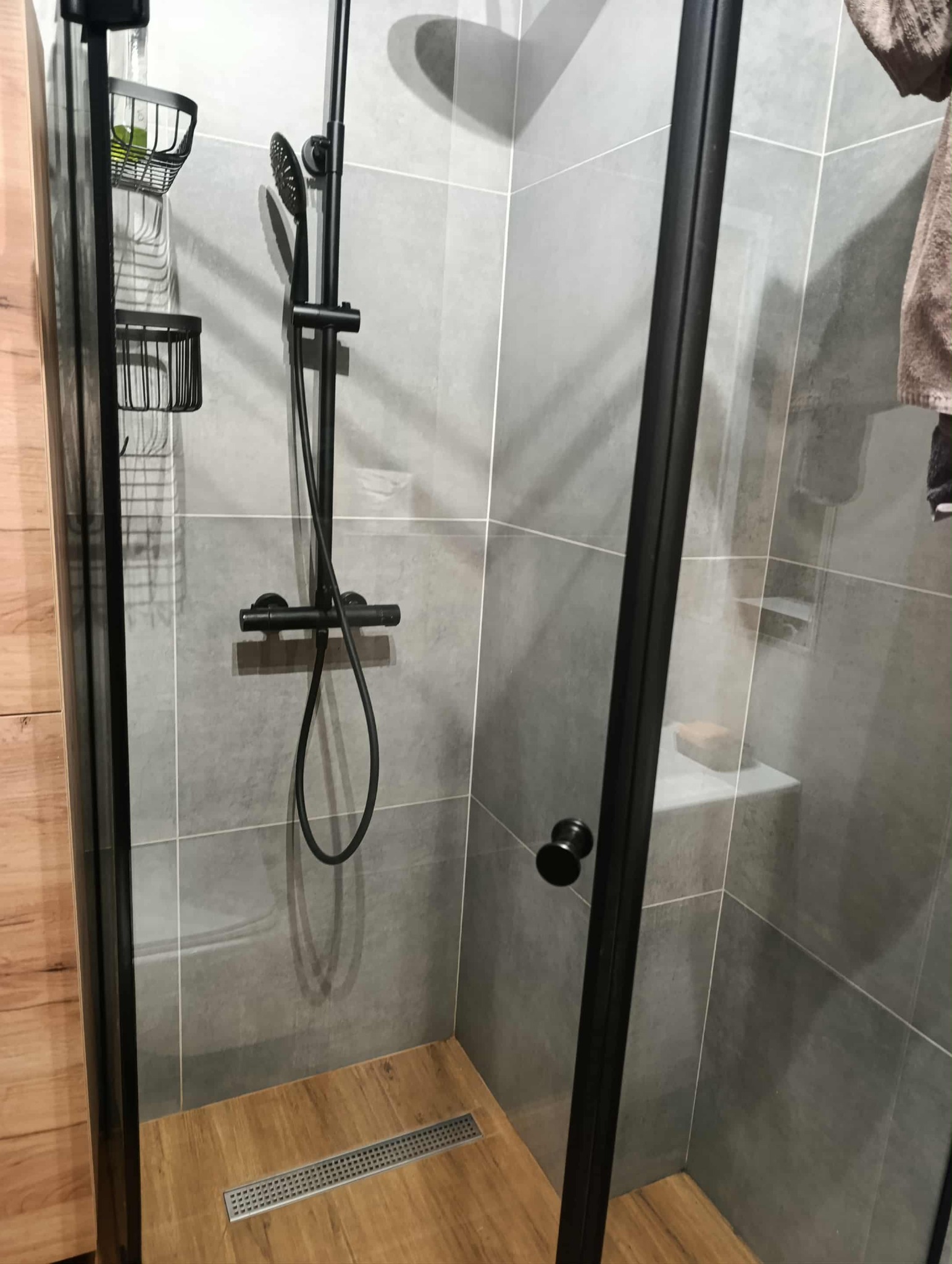 Nowoczesna kabina prysznicowa z czarnymi elementami, szare płytki i drewnopodobna podłoga z odpływem liniowym. Minimalistyczny design, widok z wnętrza kabiny.