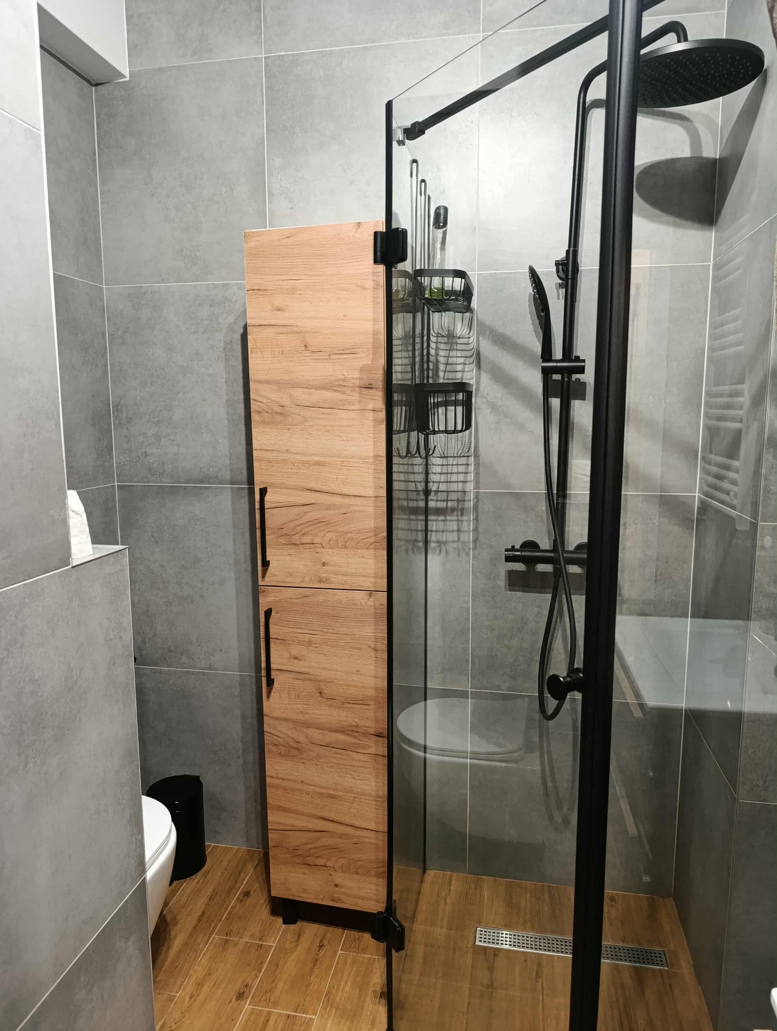 Nowoczesna łazienka po remoncie: kabina prysznicowa z czarną armaturą, drewniana szafka i szare płytki. Widoczny odpływ liniowy.