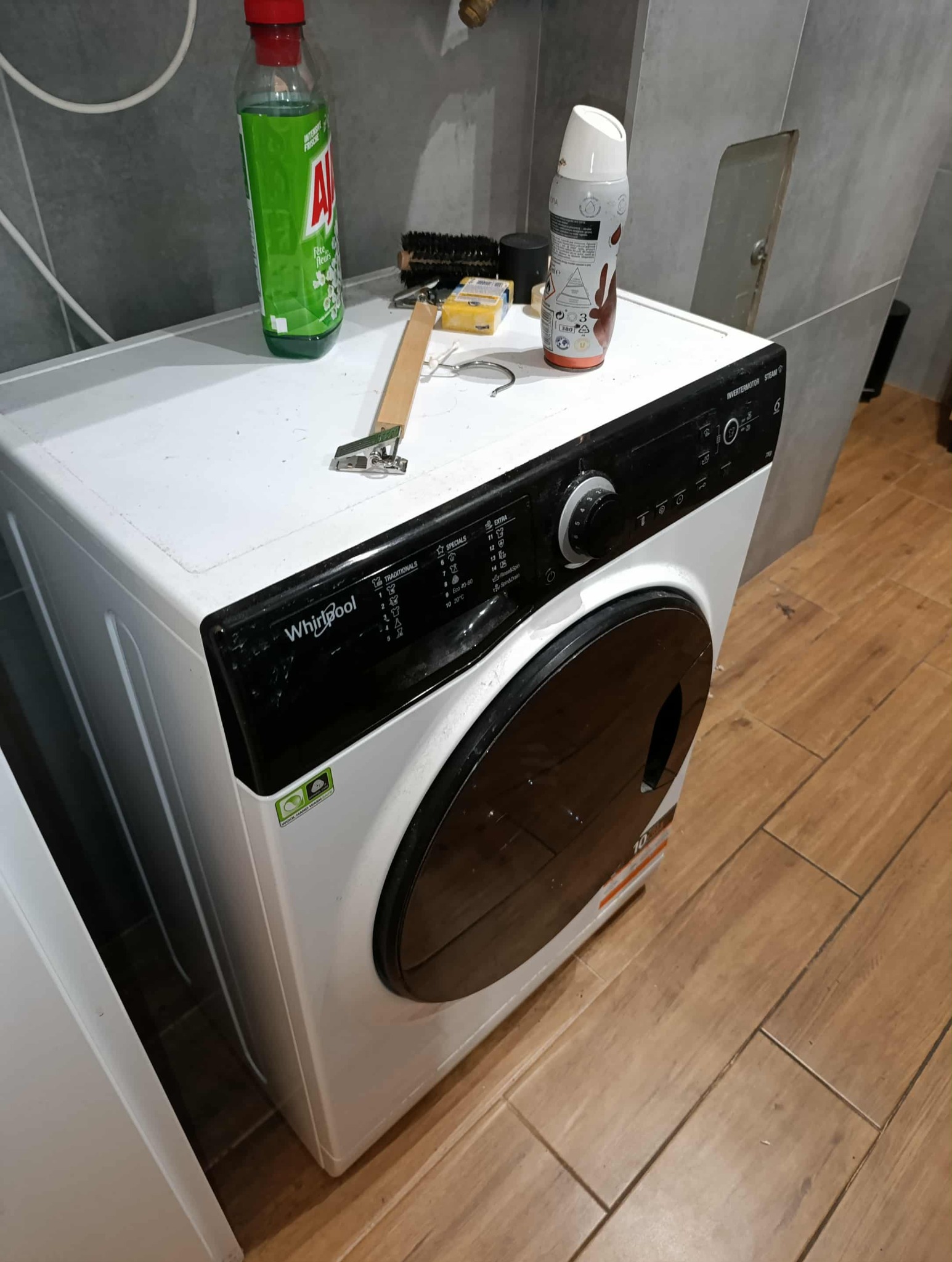 Używana pralka Whirlpool z widocznymi detergentami i narzędziami na blacie w łazience z szarymi kafelkami i drewnopodobną podłogą. Sprzęt czeka na sprzątanie.