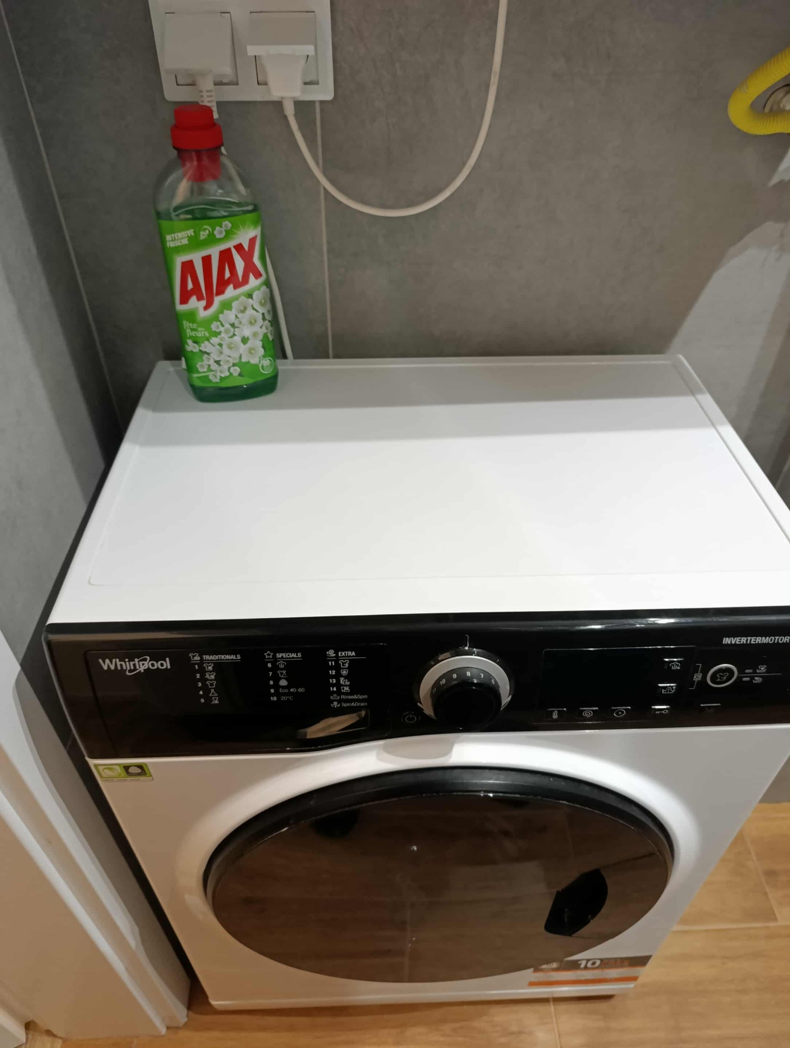 Nowoczesna pralka Whirlpool w łazience z butelką płynu Ajax na górze, gotowa do użycia. Widoczny panel sterowania i szara płytka w tle.