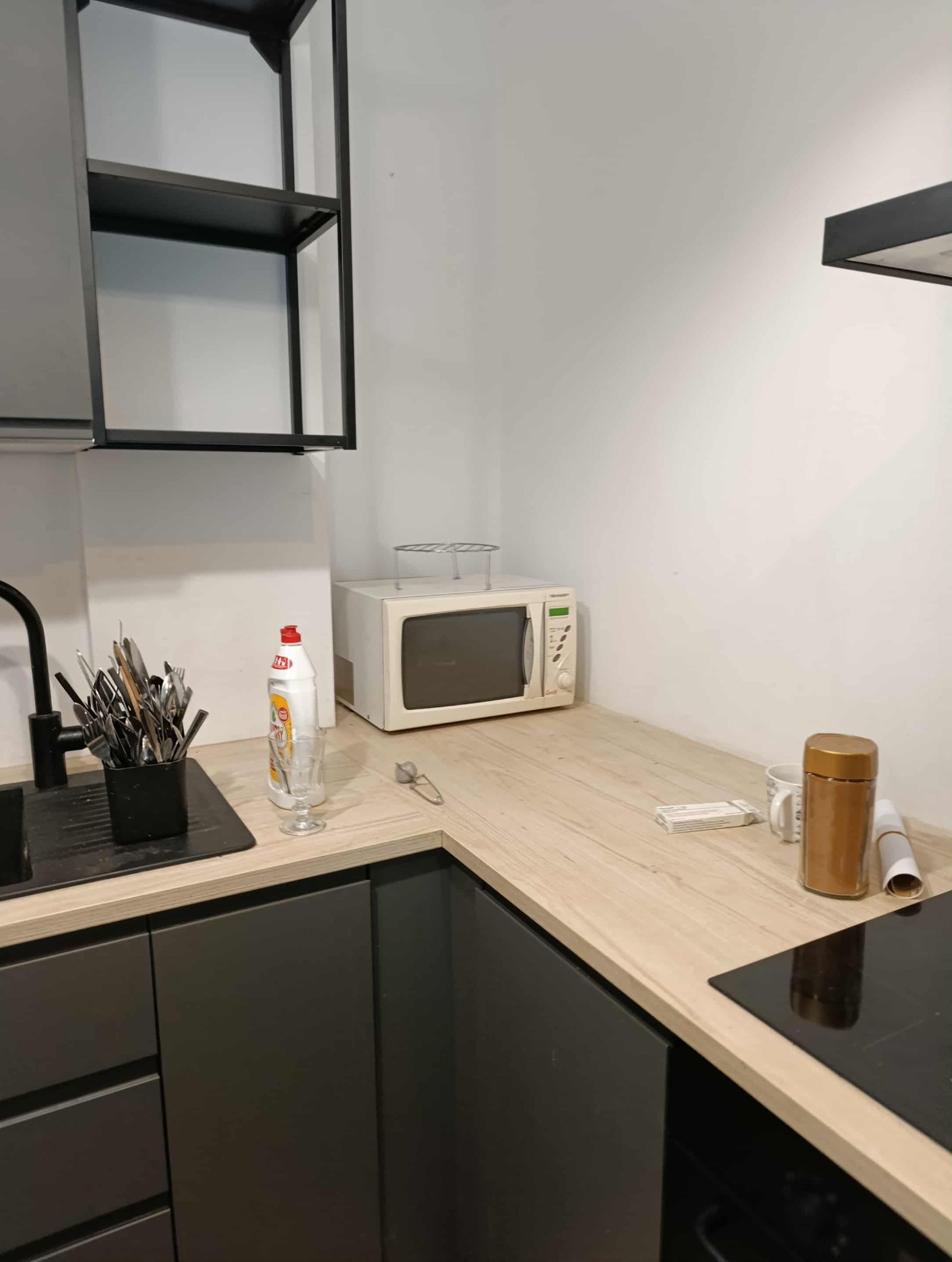 Umywalka z czarną armaturą, płyn do naczyń, szklanka i mikrofalówka na blacie w kuchni z ciemnymi szafkami. Minimalistyczny wystrój.